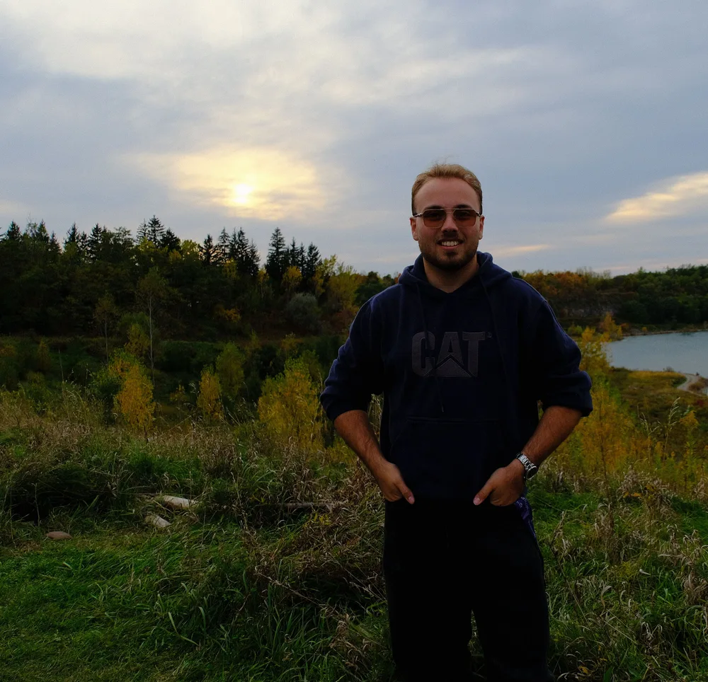 Hamza standing in tall autumn grass at Rattlesnake Point, a quarry lake and the evening sky behind him