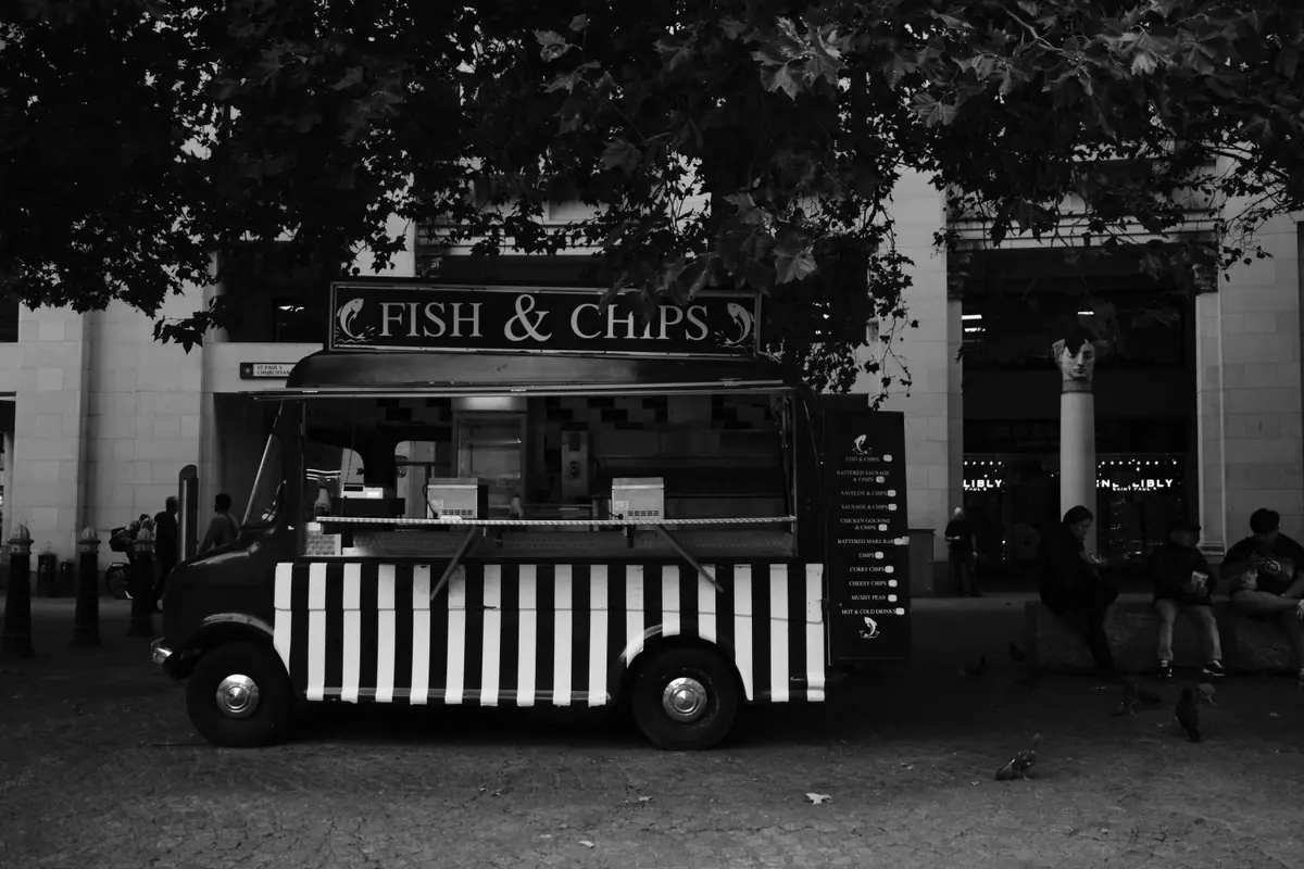 A striped fish and chips van parked under trees, in black and white