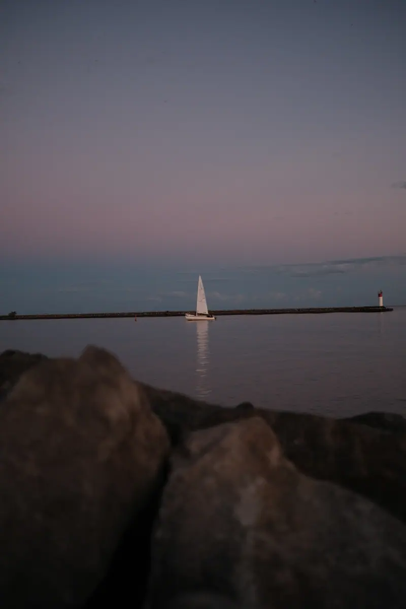 A sailboat passes the pier at dusk, rocks in the foreground