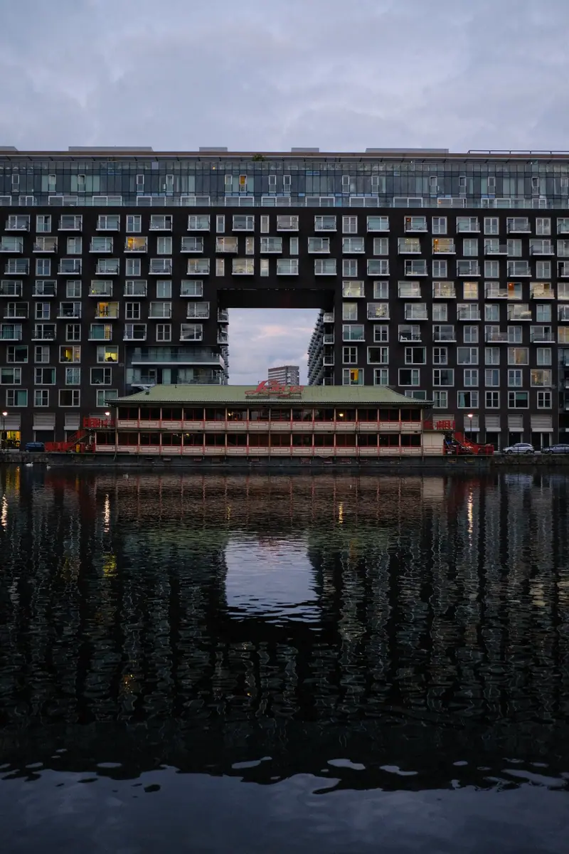 A large building with a square opening reflected in dark dock water, a floating restaurant beneath it
