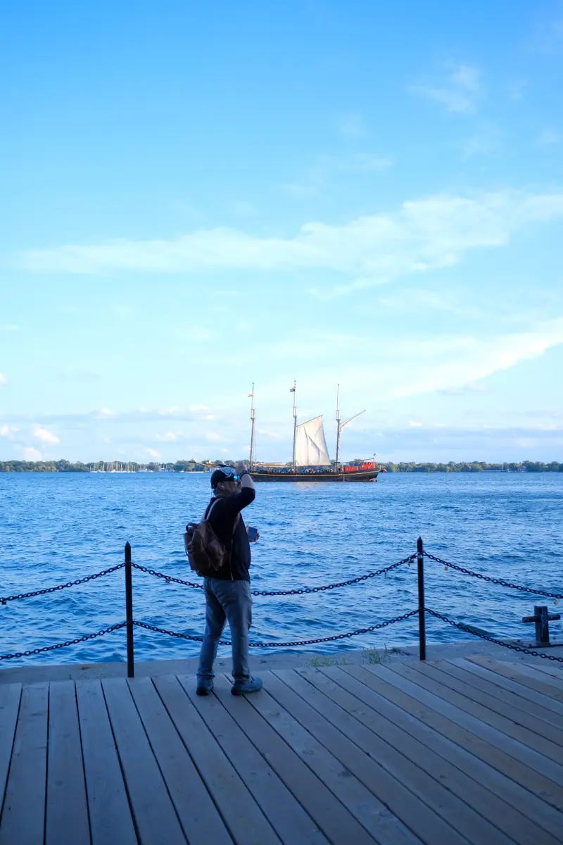A man in a cap stands at the chain railing of a boardwalk on Toronto's waterfront, a tall ship under sail on the lake behind him
