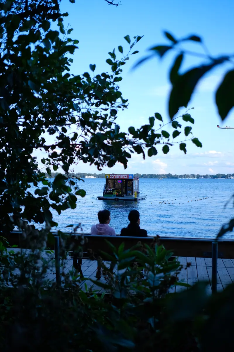 Two people on a bench watch a small floating convenience store cross the harbour, framed by dark leaves