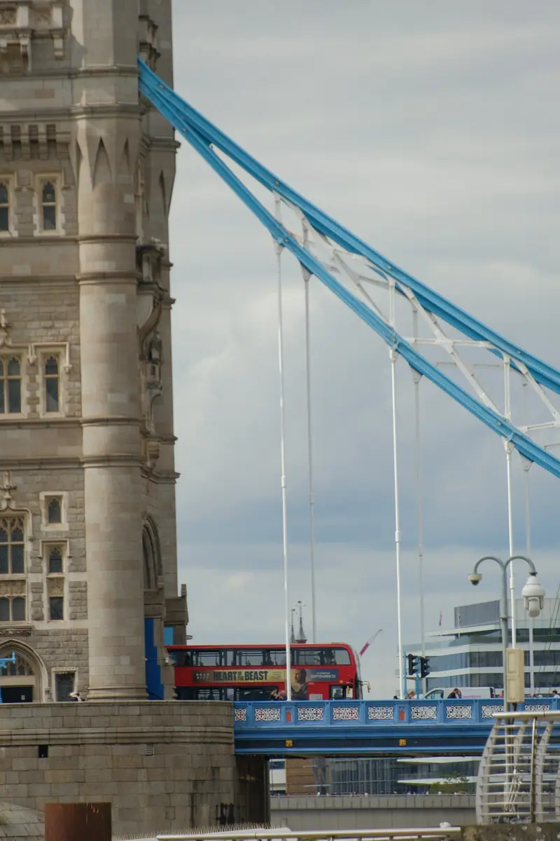 A tower of Tower Bridge and its blue suspension chains, a red bus crossing below