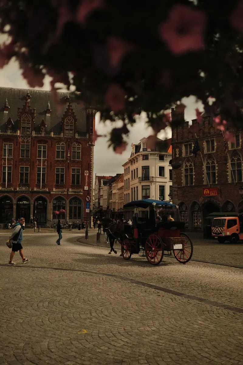 A horse-drawn carriage crosses the market square in Bruges, framed by pink flowers overhead