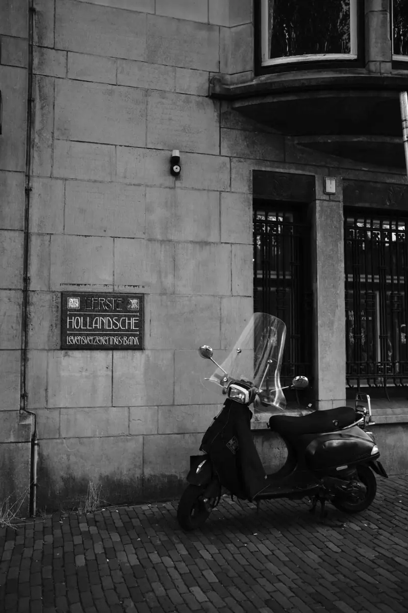 A black scooter parked under a stone Hollandsche building sign, in black and white