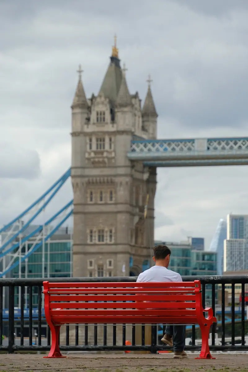 A man sits on a red bench facing Tower Bridge
