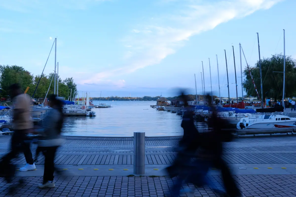 People blur past along the harbourfront promenade, sailboats moored behind them