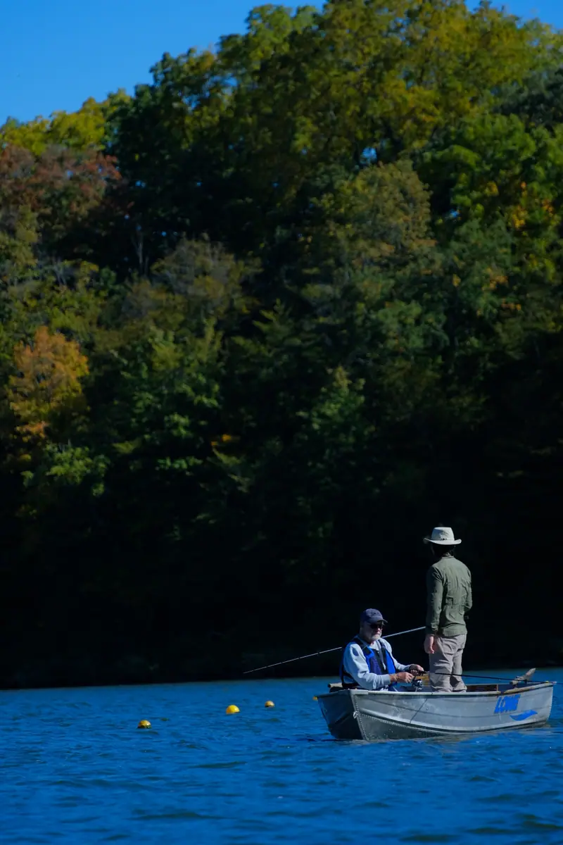Two people fishing from a small aluminium boat below autumn trees