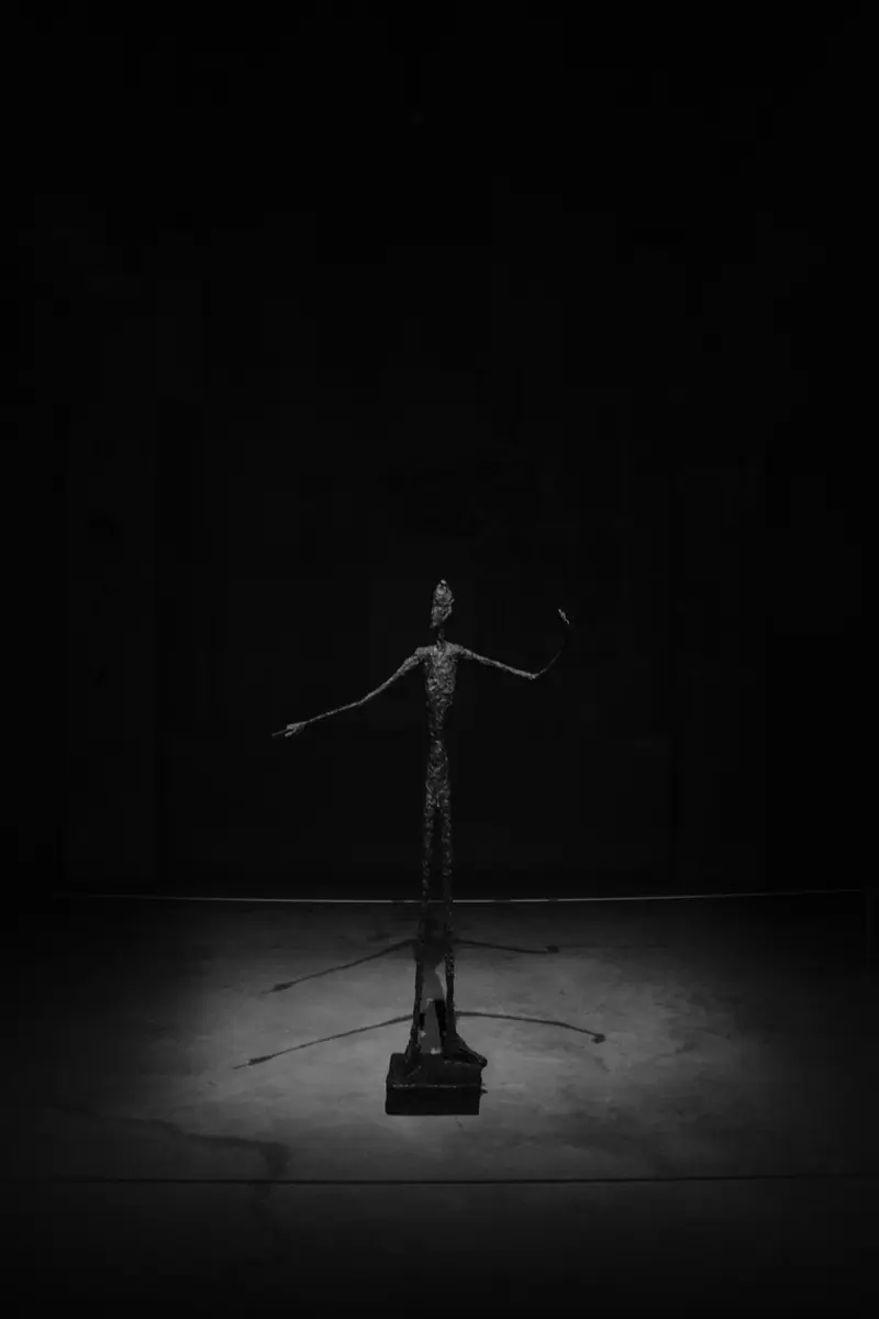 A thin bronze figure with outstretched arms under a single spotlight in a dark gallery