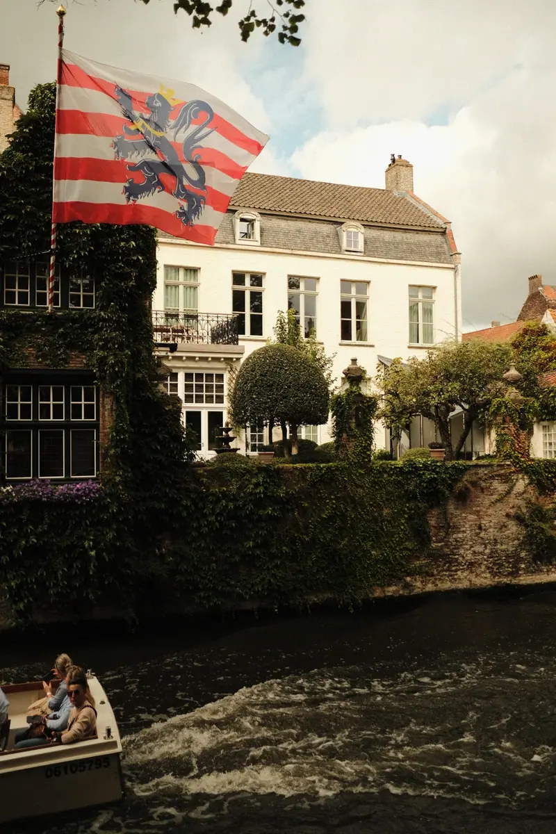 The red-and-white striped flag of Bruges flies above a canal house as a tour boat passes