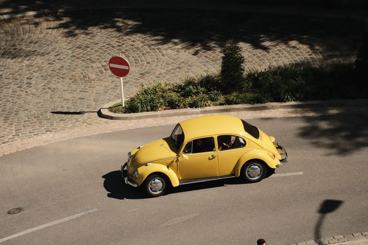A yellow Volkswagen Beetle rounds a curve on a cobbled road, seen from above